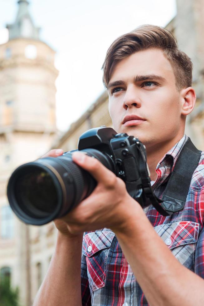 fiducioso fotografo. bello giovane uomo Tenere digitale telecamera e guardare lontano mentre in piedi all'aperto foto