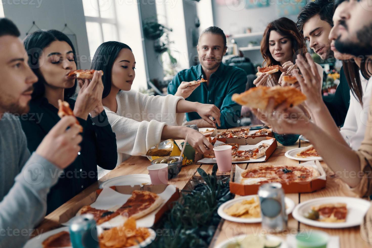 così delizioso gruppo di giovane persone nel casuale indossare mangiare Pizza e sorridente mentre avendo un' cena festa in casa foto