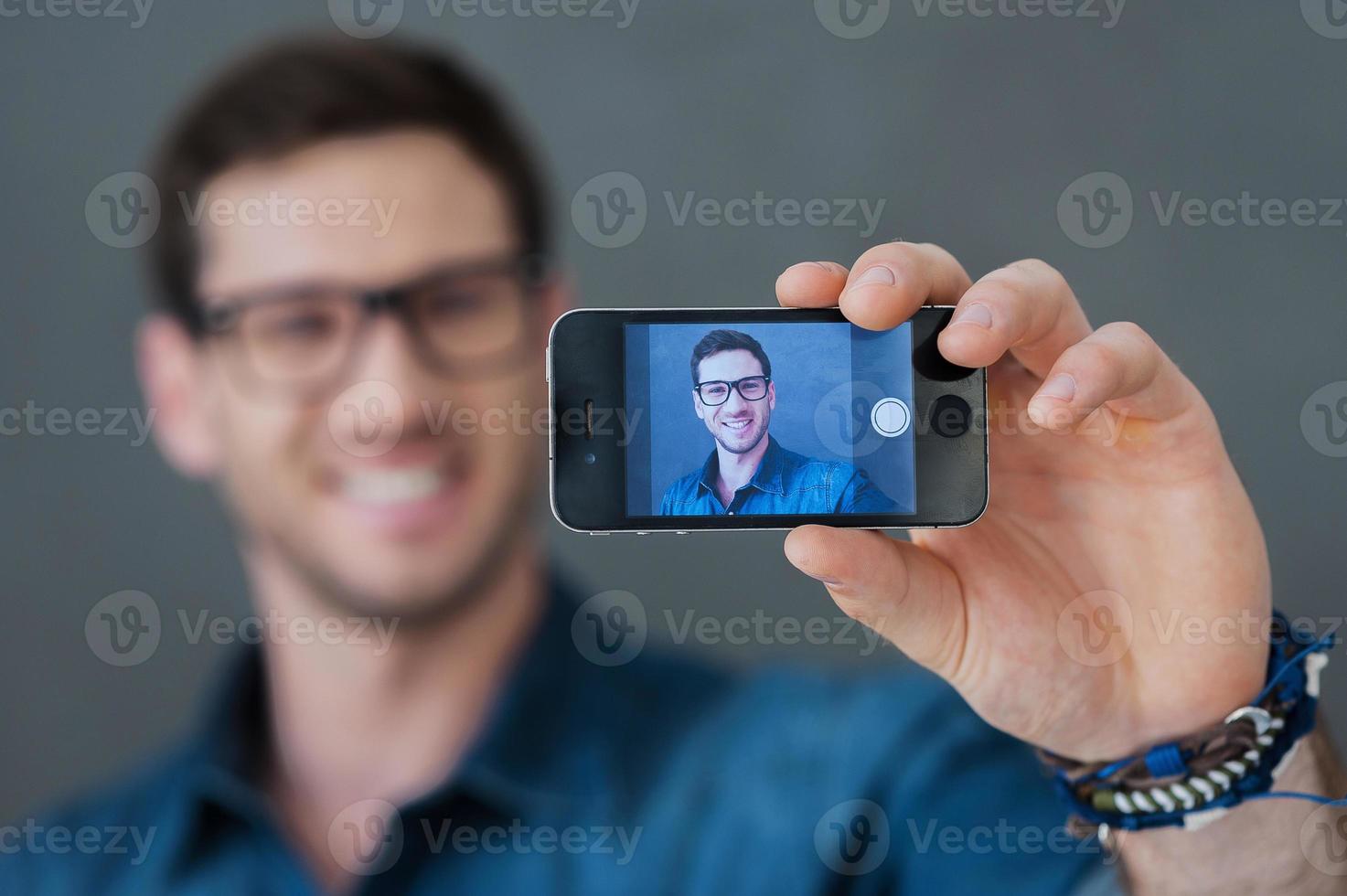 fabbricazione contento autoscatto. sorridente giovane uomo fabbricazione autoscatto di il suo inteligente Telefono mentre in piedi contro grigio sfondo foto
