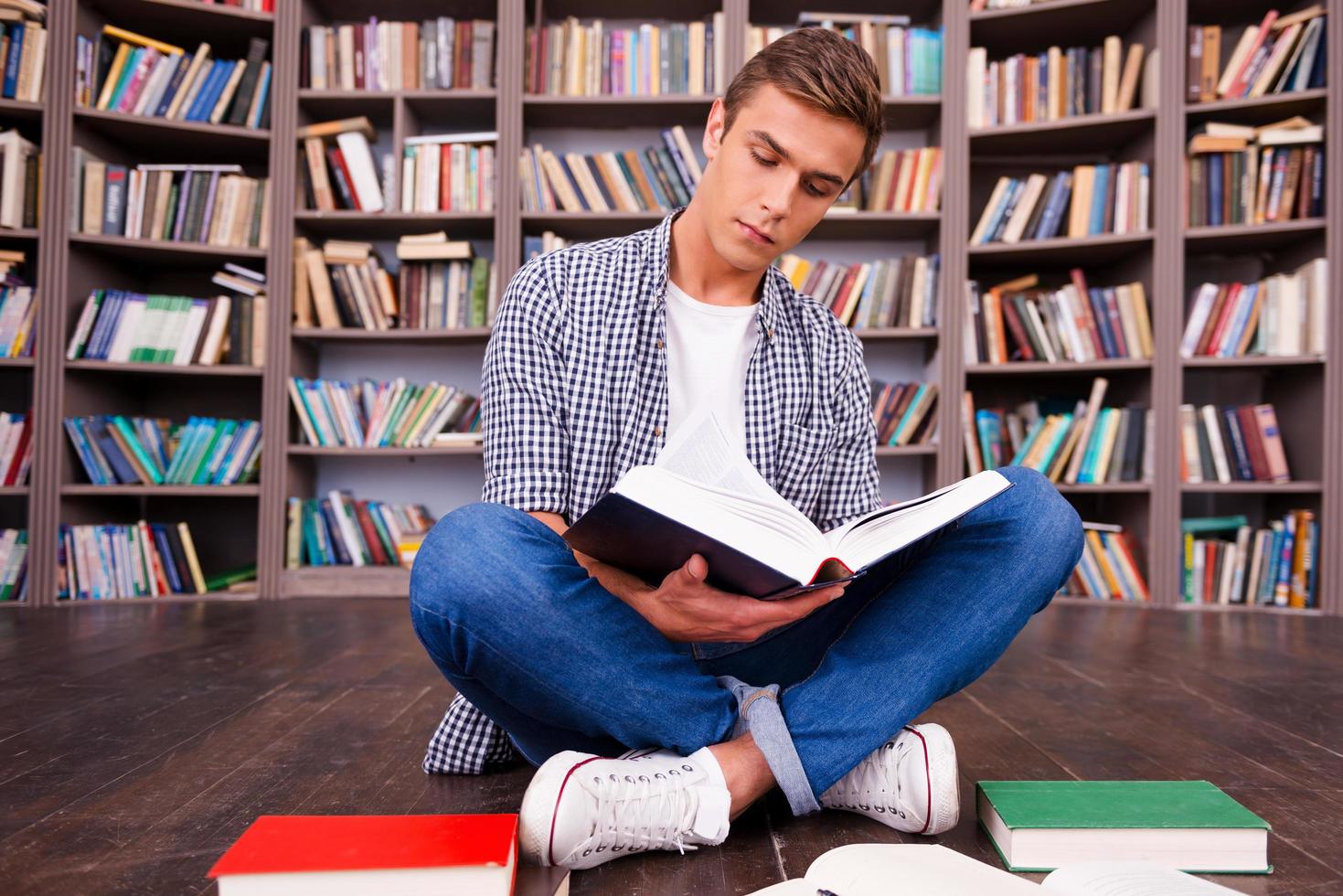 fare il suo ricerca nel biblioteca. concentrato giovane uomo lettura libro mentre seduta contro scaffale foto