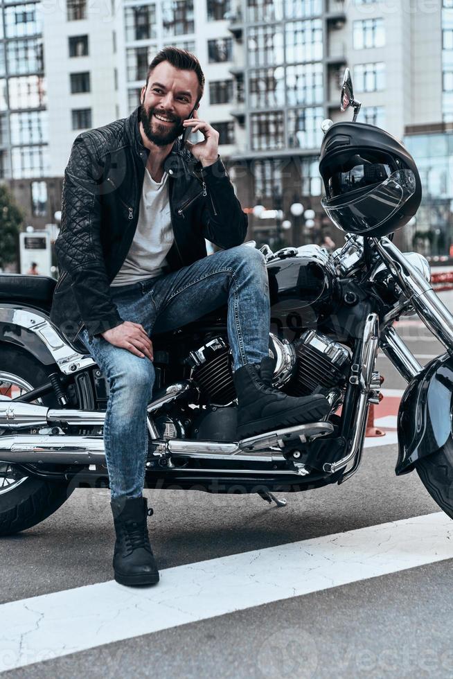 lieto per sentire voi bello giovane uomo nel pelle giacca parlando su il Telefono e sorridente mentre seduta su il motocicletta all'aperto foto