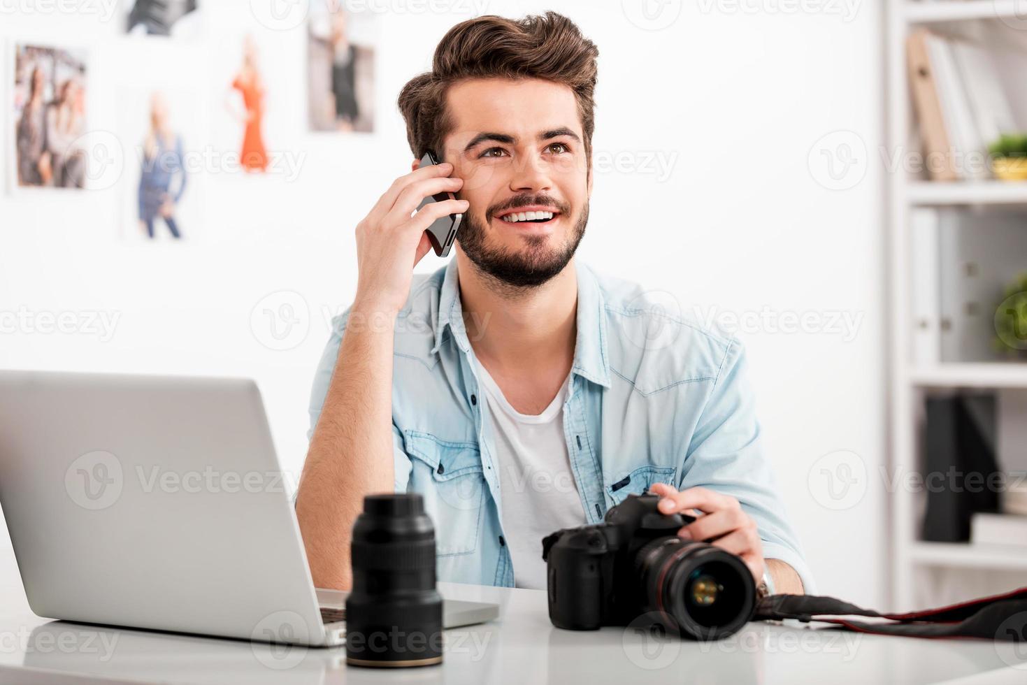 godendo creativo opera. contento giovane uomo parlando su il mobile Telefono e sorridente mentre seduta a il suo Lavorando posto e Tenere digitale telecamera foto