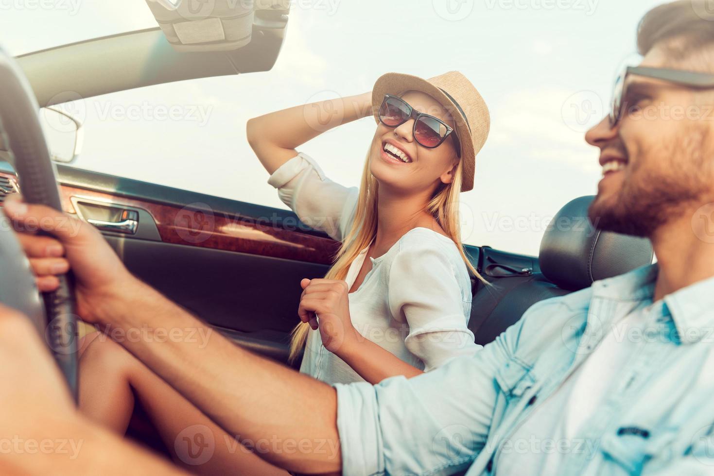 avendo divertimento lungo il modo. bellissimo giovane donna sorridente e regolazione sua cappello mentre sua fidanzato seduta vicino e guida convertibile foto