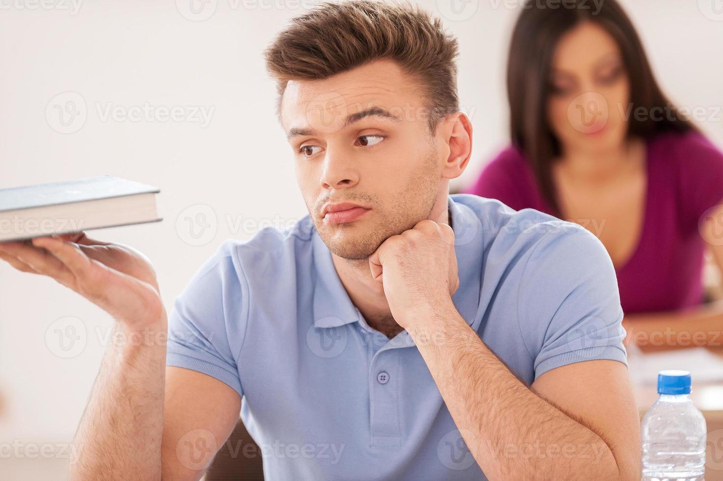 annoiato alunno. bello maschio alunno Tenere mano su mento e guardare a il manuale mentre giovane donna seduta a il scrivania dietro a lui foto