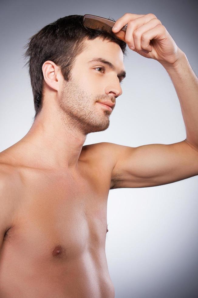 uomo pettinatura capelli. lato Visualizza di bello giovane barba uomo guardare a telecamera e pettinatura il suo capelli mentre in piedi isolato su grigio sfondo foto