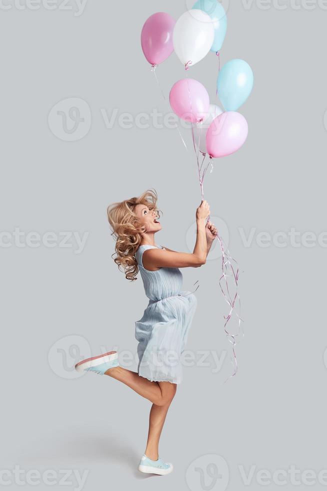 pronto per volare alto. pieno lunghezza studio tiro di giocoso giovane donna Tenere palloncini e sorridente mentre salto contro grigio sfondo foto