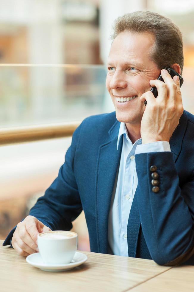 soggiorno collegato. fiducioso maturo uomo nel vestito formale potabile caffè e parlando su mobile Telefono mentre seduta nel ristorante foto