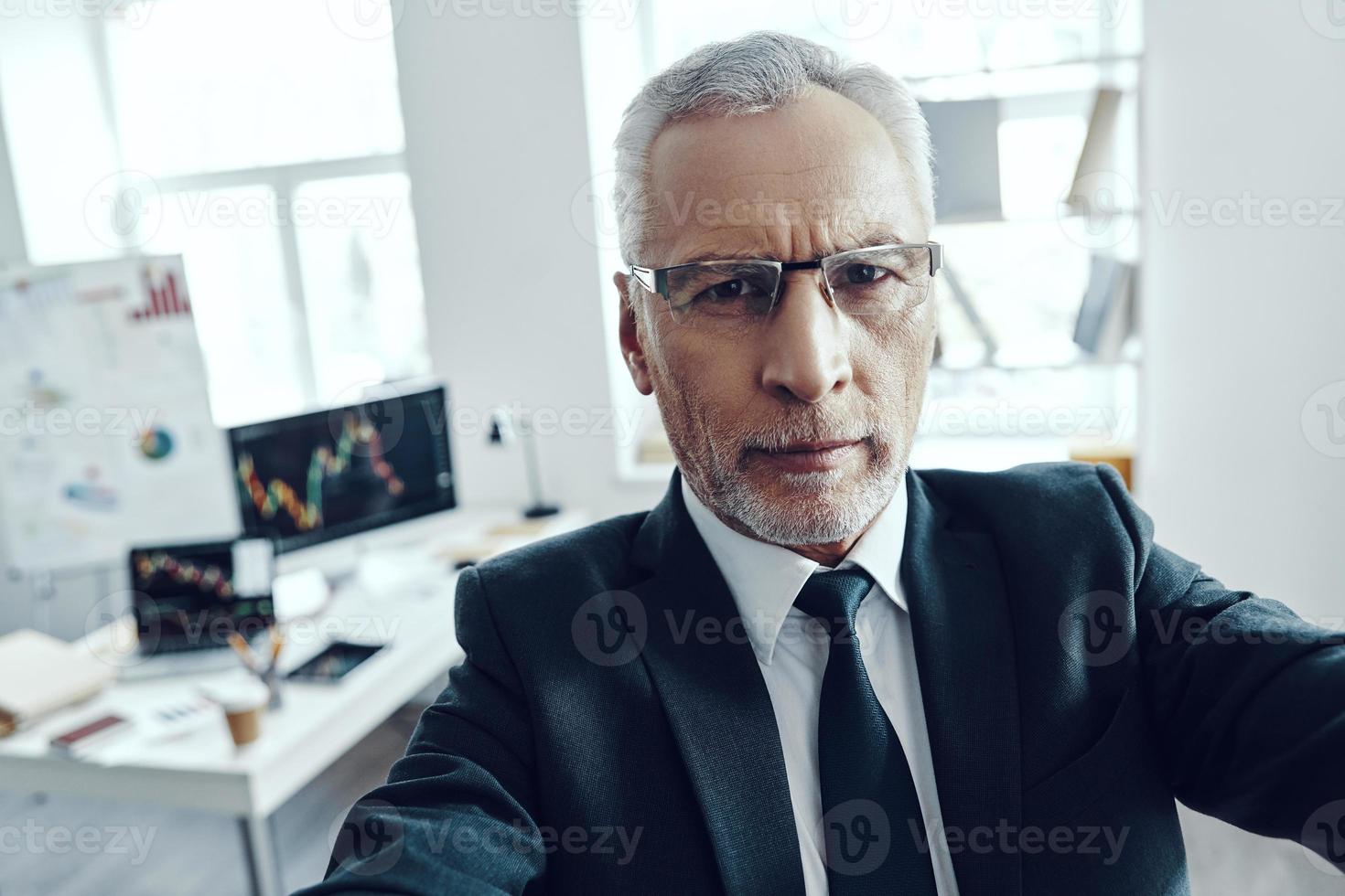 se stesso ritratto di anziano uomo nel elegante attività commerciale completo da uomo guardare a telecamera mentre in piedi nel ufficio foto