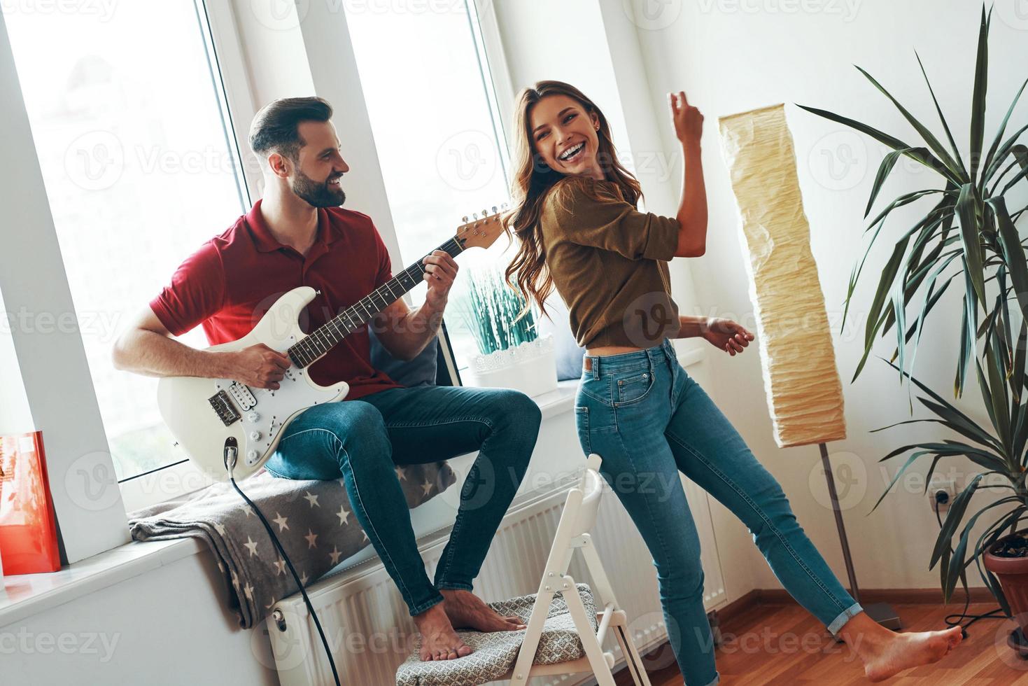 contento giovane uomo nel casuale capi di abbigliamento giocando il chitarra mentre il suo attraente fidanzata danza foto