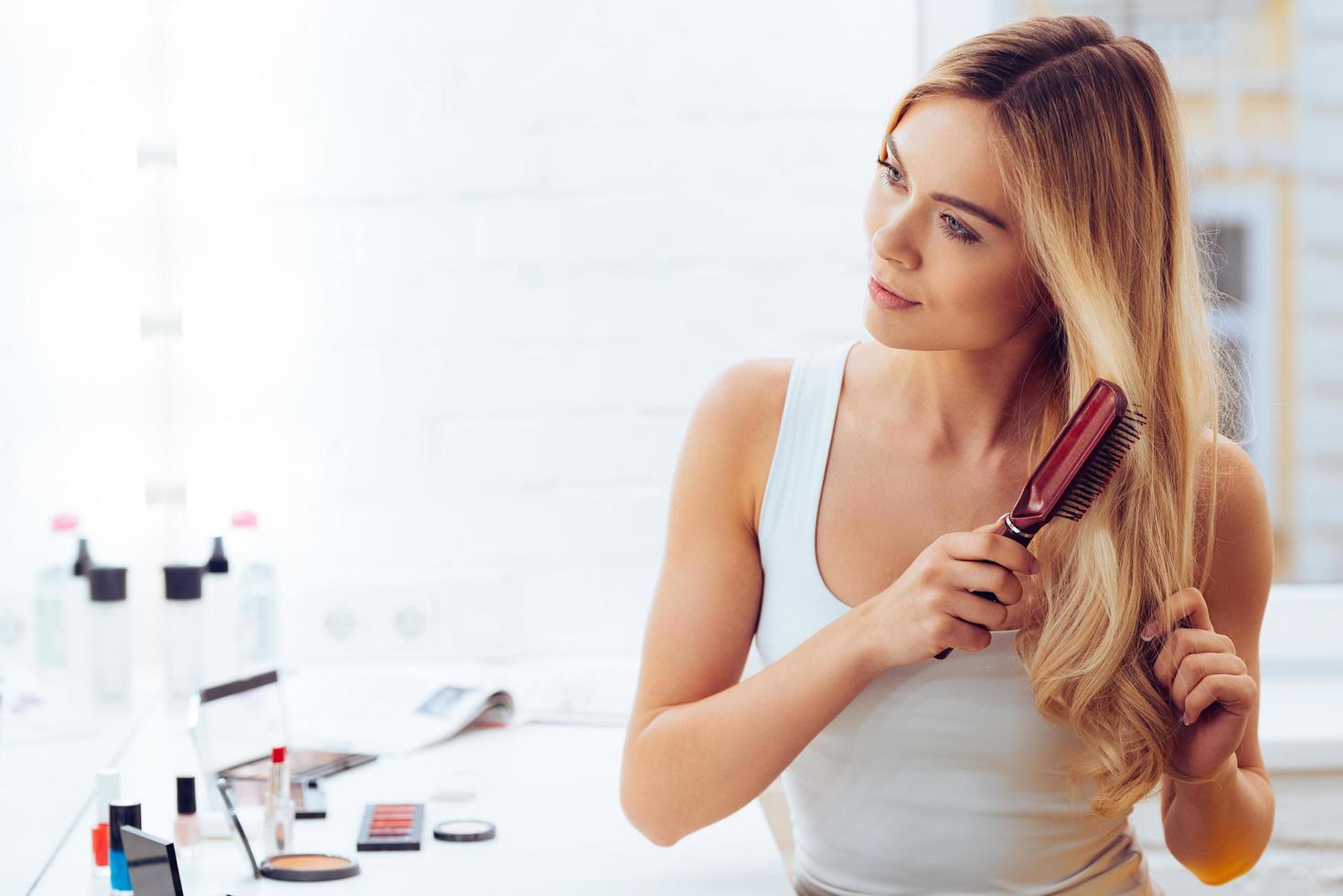 ottenere sbarazzarsi di grovigli. bellissimo giovane donna guardare a sua riflessione nel mirrorand spazzolatura sua lungo capelli mentre seduta a il condimento tavolo foto