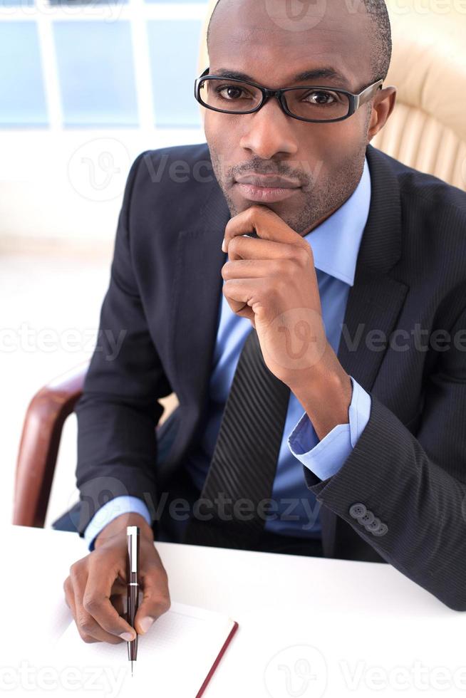 riflessivo uomo d'affari. superiore Visualizza di giovane africano uomo d'affari scrittura qualcosa nel il suo Nota tampone e Tenere mano su mento mentre seduta a il suo Lavorando posto foto