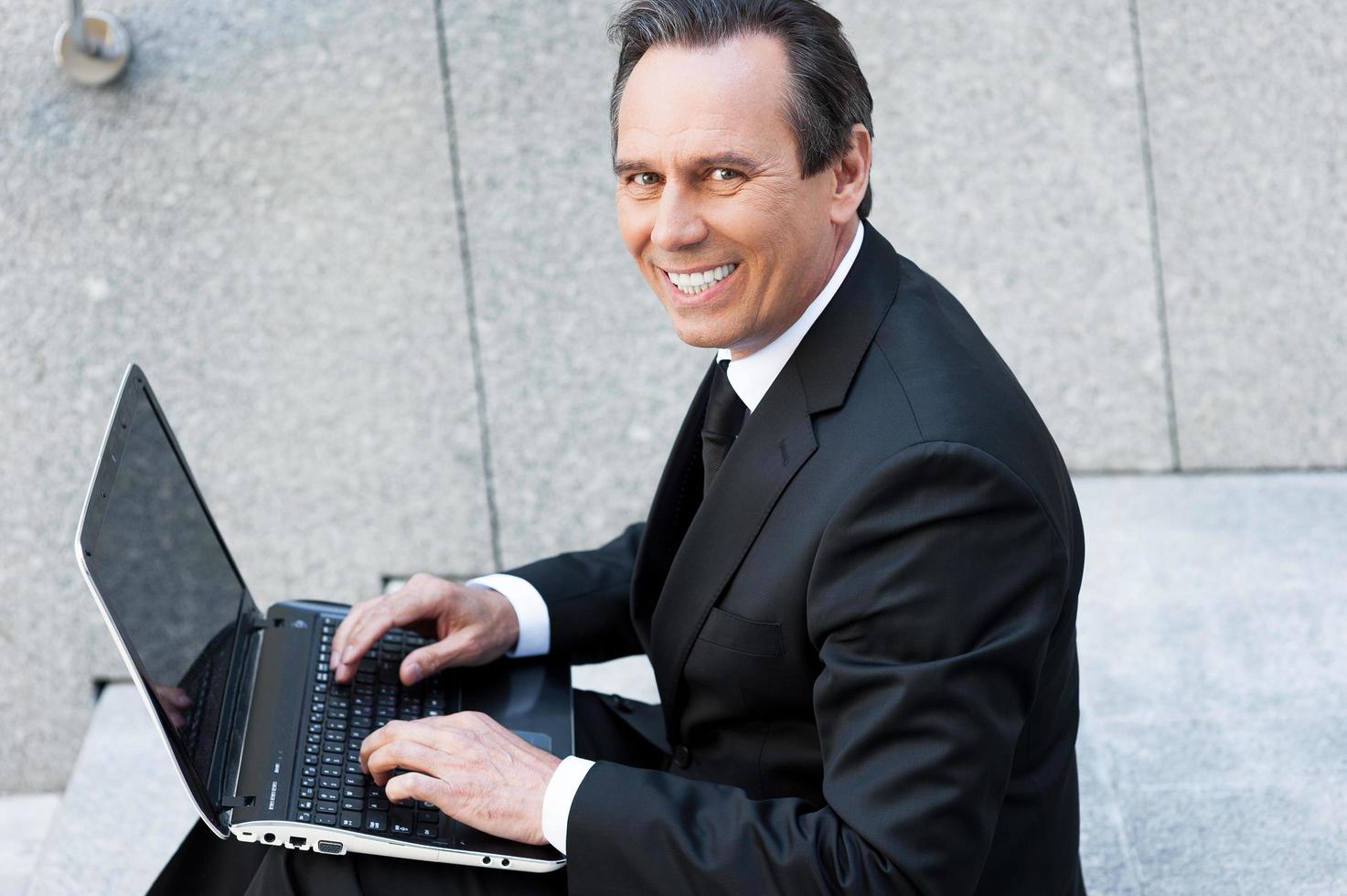 fiducioso uomo d'affari. allegro anziano uomo nel vestito formale Lavorando su il computer portatile e sorridente mentre seduta all'aperto foto
