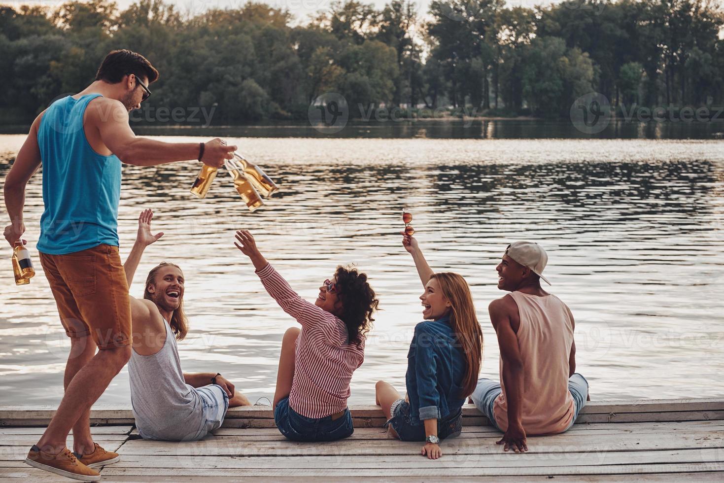 godendo spensierato volta. gruppo di contento giovane persone nel casuale indossare sorridente e potabile birra mentre seduta su il molo foto