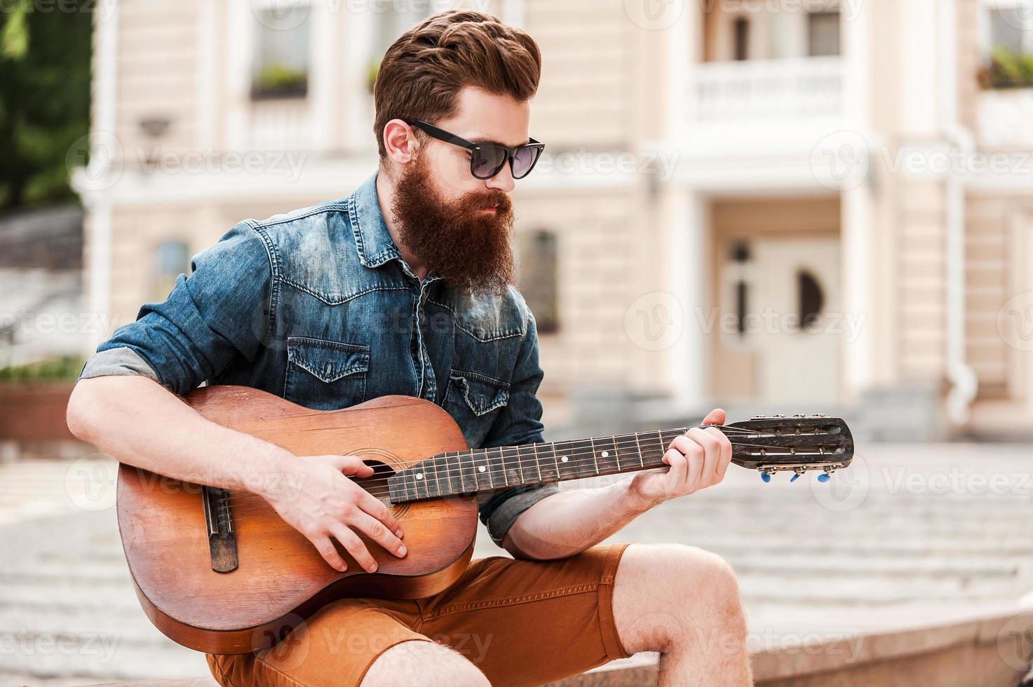 strada musicista. bello giovane barbuto uomo giocando il chitarra mentre seduta all'aperto foto