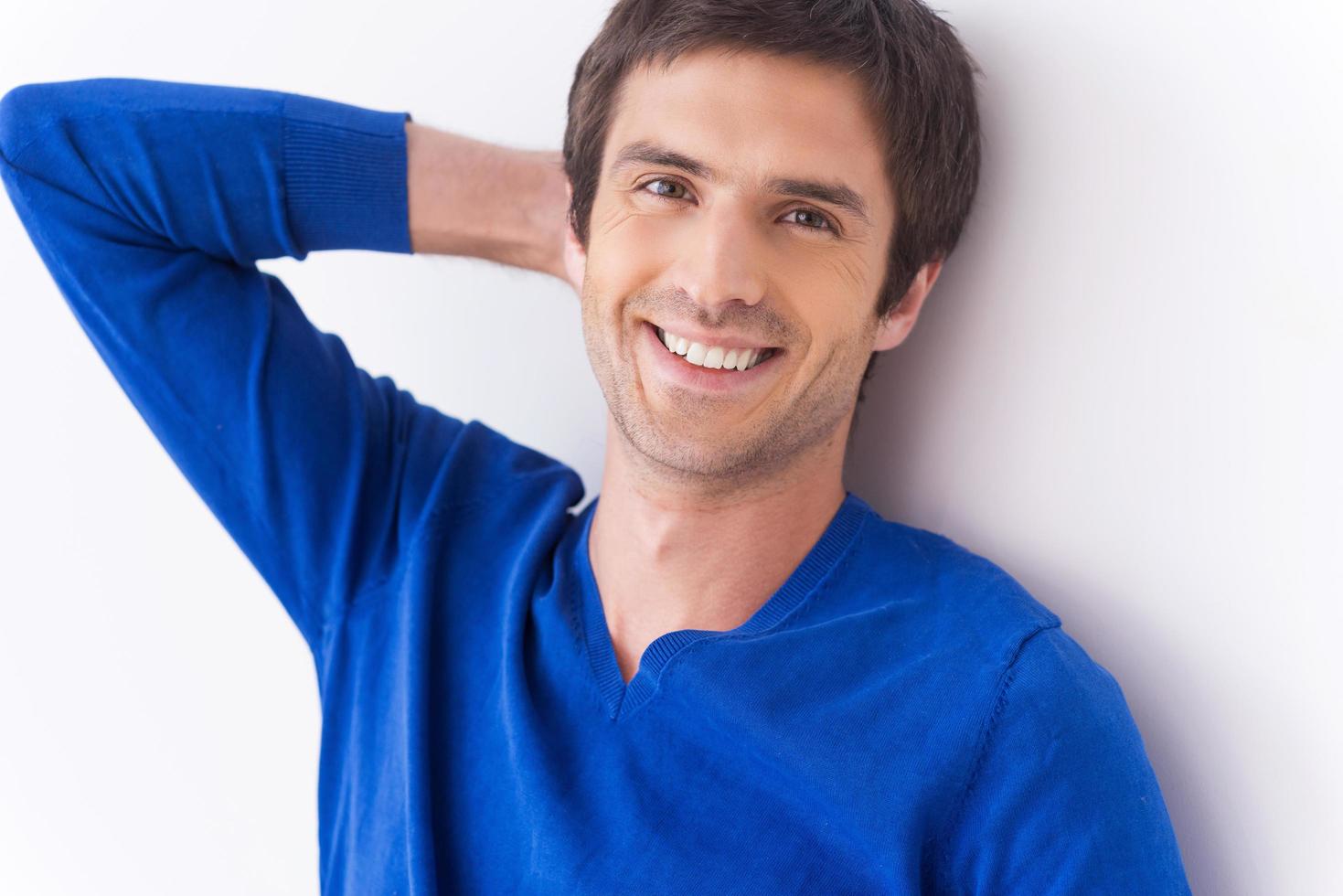 bello e sicuro di sé. bello giovane uomo nel blu maglione guardare a telecamera e sorridente mentre Tenere mano dietro a testa e in piedi contro grigio sfondo foto