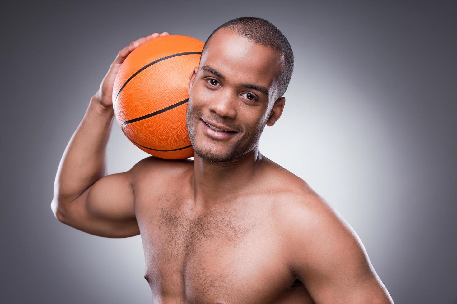 pronto per un' gioco. giovane A petto nudo africano uomo Tenere pallacanestro palla e guardare a telecamera mentre in piedi contro grigio sfondo foto