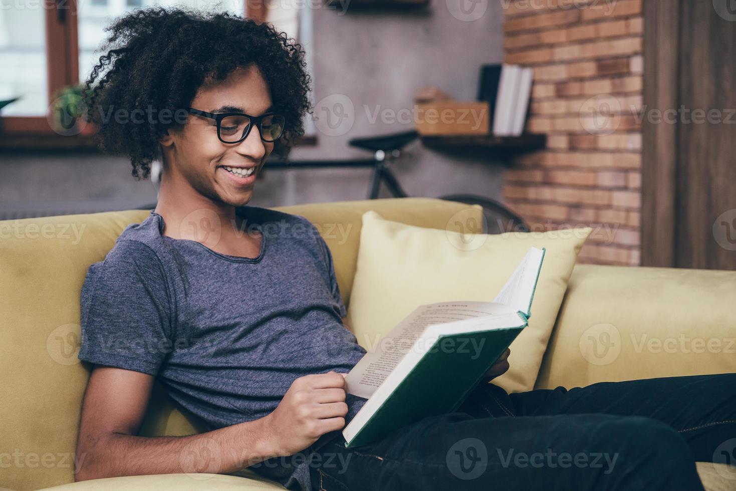 godendo nuovo capitolo. lato Visualizza di allegro giovane africano uomo lettura libro con Sorridi e indossare bicchieri mentre seduta su il divano a casa foto