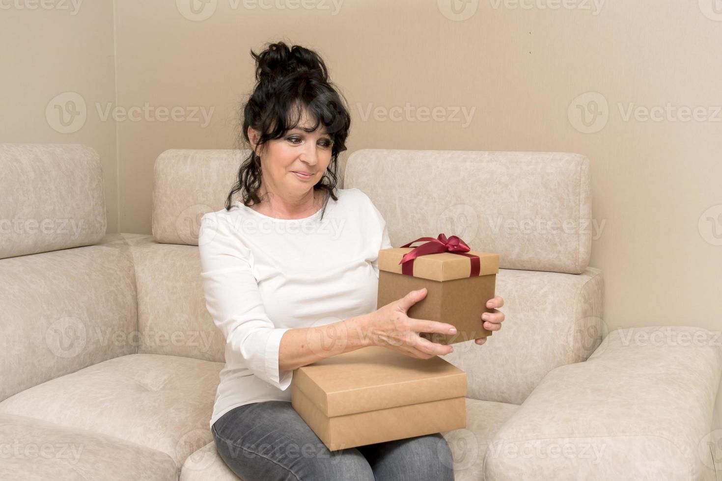 bellissimo anziano donna seduta su il divano con piacere esamina il ricevuto i regali foto