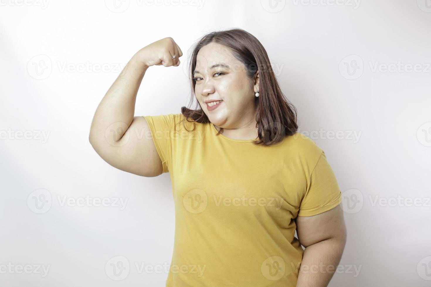 eccitato asiatico grande dimensioni donna indossare un' giallo maglietta mostrando forte gesto di sollevamento sua braccia e muscoli sorridente con orgoglio foto