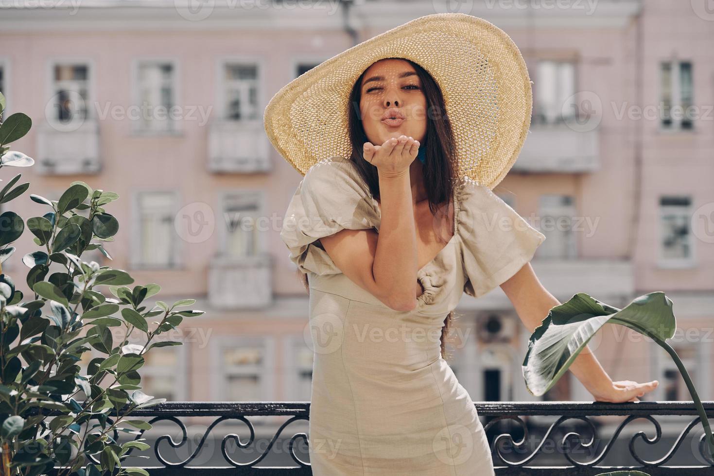 pieno lunghezza di bellissimo giovane donna nel elegante cappello soffiaggio un' bacio mentre rilassante su il balcone foto