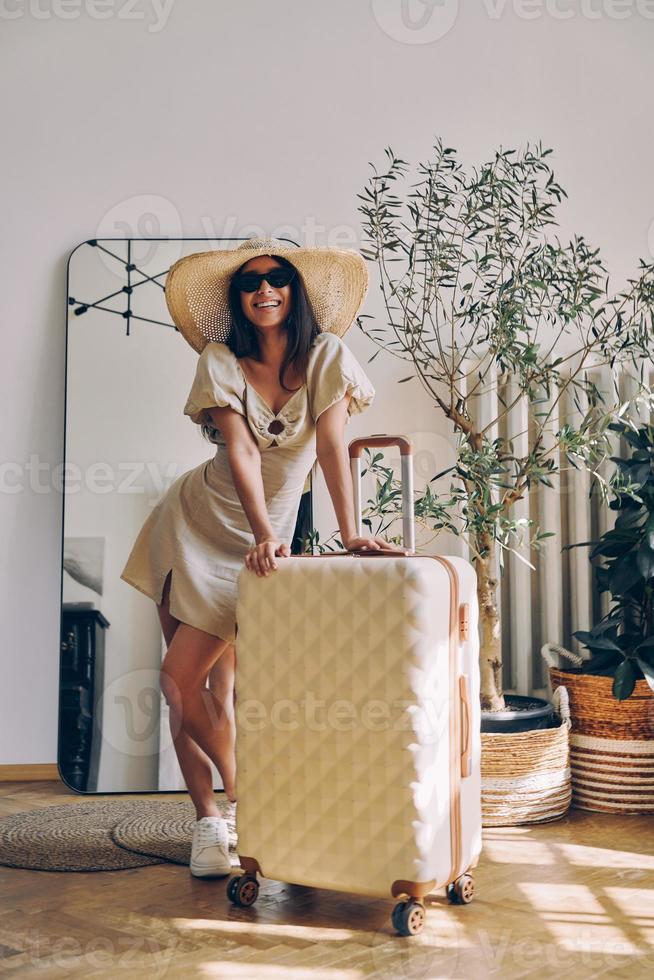 bellissimo giovane donna nel elegante cappello pendente su il valigia e sorridente mentre in piedi a casa foto