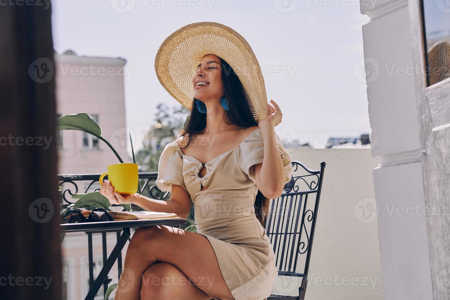 gioioso giovane donna nel elegante cappello Tenere caffè tazza e sorridente mentre rilassante su il balcone foto