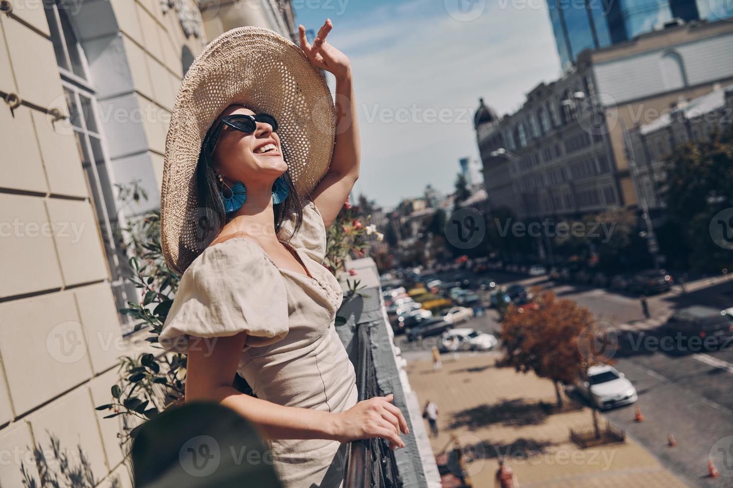 rilassato giovane donna regolazione sua elegante cappello e sorridente mentre in piedi su il balcone foto