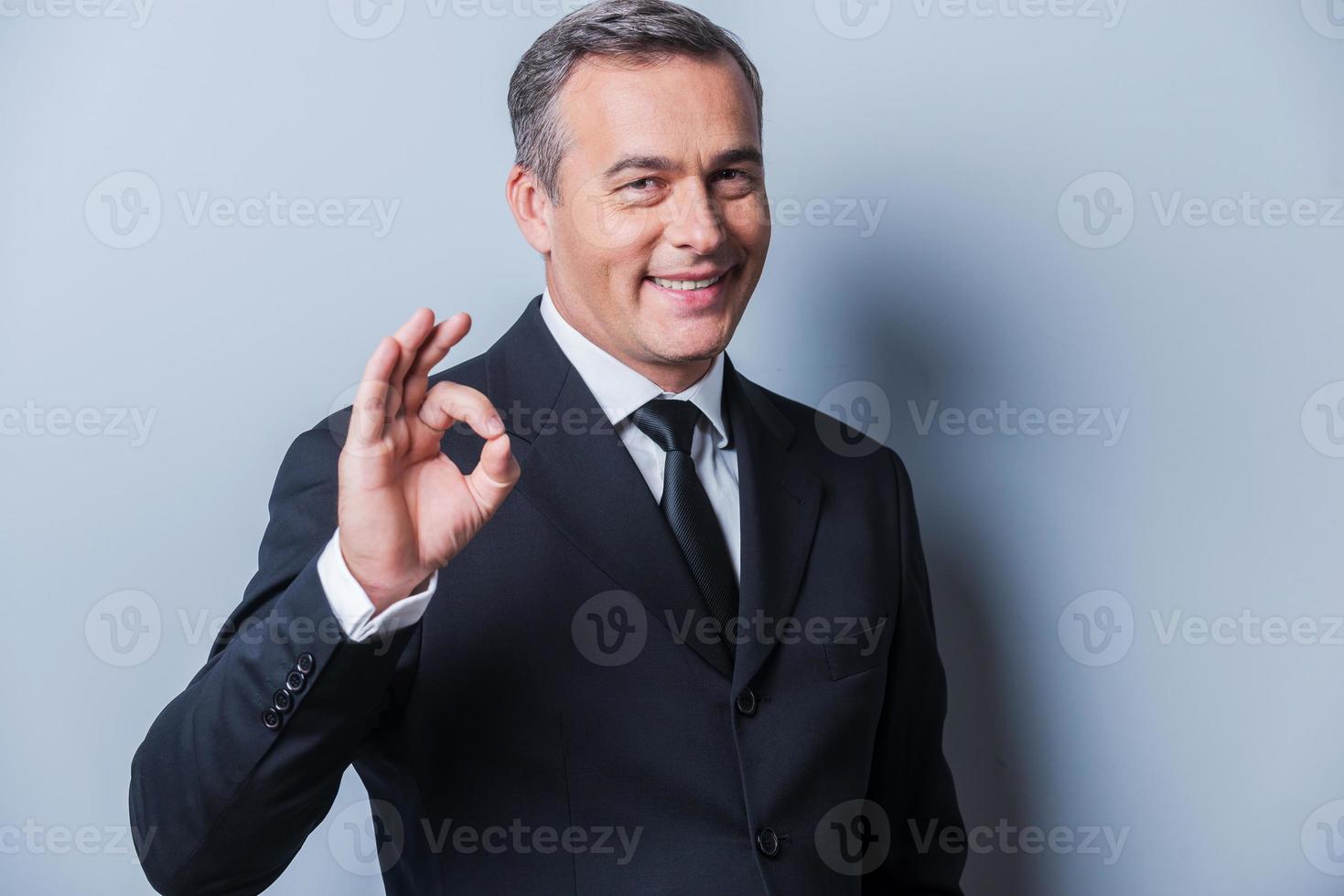qualunque cosa è ok allegro maturo uomo nel vestito formale Esprimere a gesti ok cartello e sorridente mentre in piedi contro grigio sfondo foto