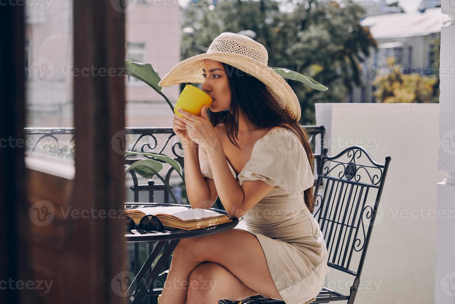 attraente giovane donna nel elegante cappello potabile caffè mentre rilassante su il balcone foto