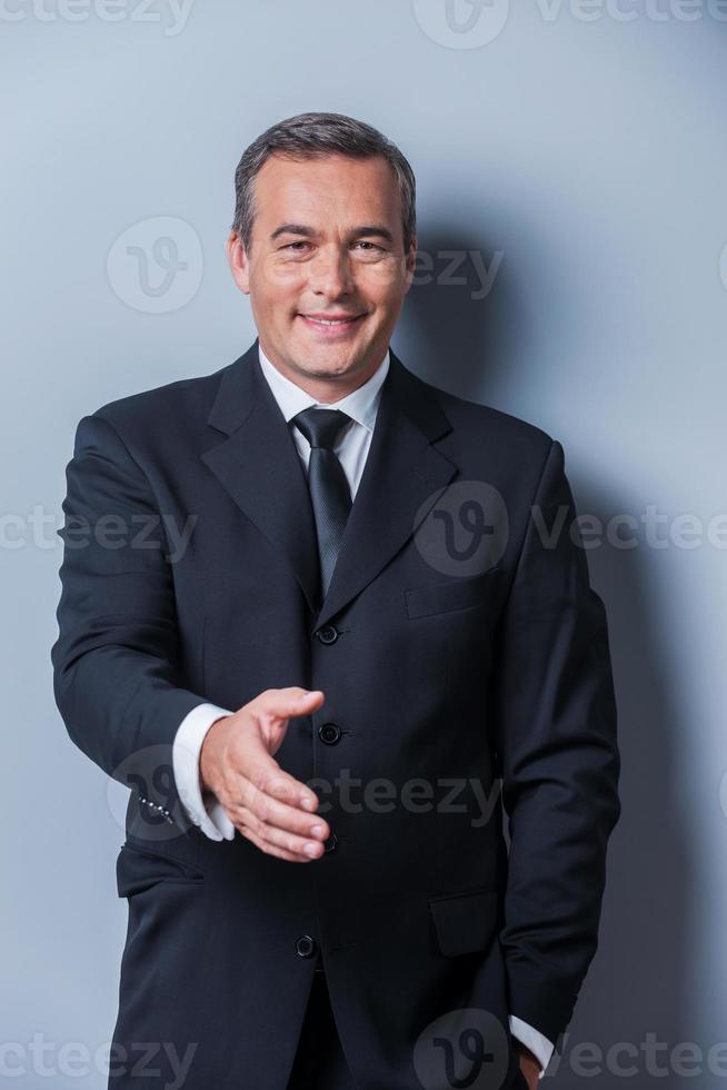 Congratulazioni ritratto di allegro maturo uomo nel vestito formale allungamento su mano per tremante mentre in piedi contro grigio sfondo foto
