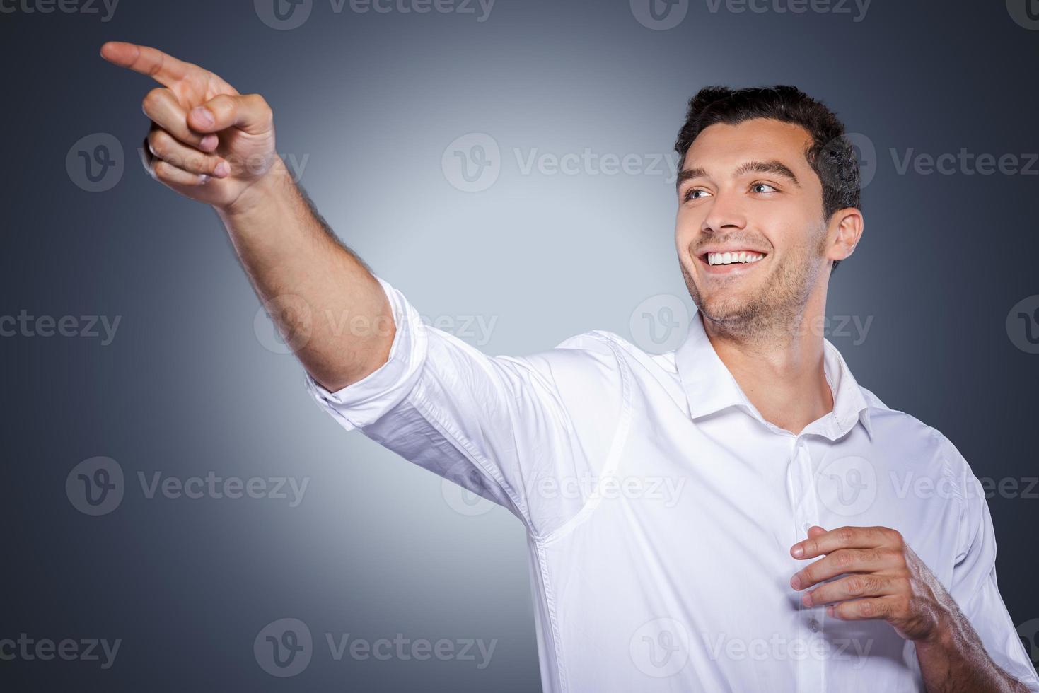 appena Guarda a quello contento giovane uomo nel bianca camicia puntamento lontano e sorridente mentre in piedi contro grigio sfondo foto