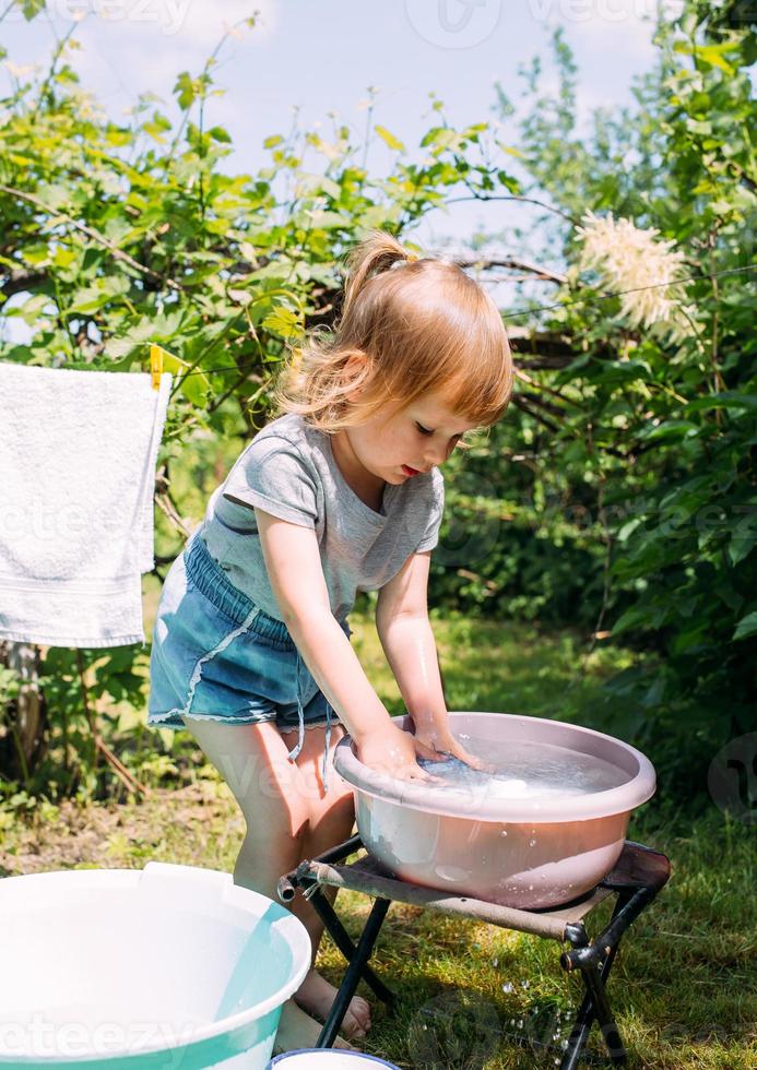 la bambina in età prescolare aiuta con il bucato. il bambino lava i vestiti in giardino foto