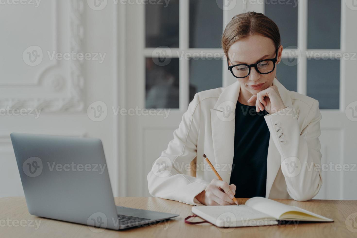 donna d'affari scrive nel taccuino, fabbricazione Appunti, pianificazione orario, seduta a ufficio scrivania con il computer portatile foto