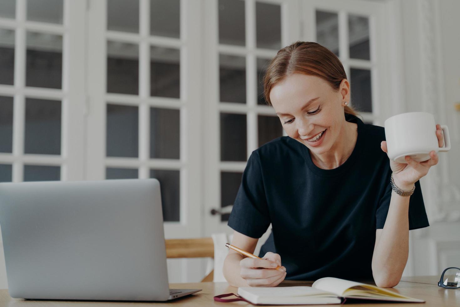 sorridente ragazza scrittura Appunti, pianificazione programma per giorno, Tenere caffè boccale su posto di lavoro con il computer portatile foto