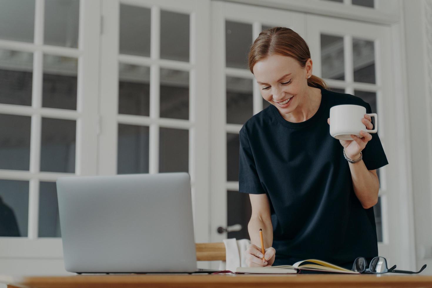 femmina scrittura Appunti nel taccuino in piedi a scrivania, Tenere caffè tazza, pianificazione programma nel posto di lavoro foto