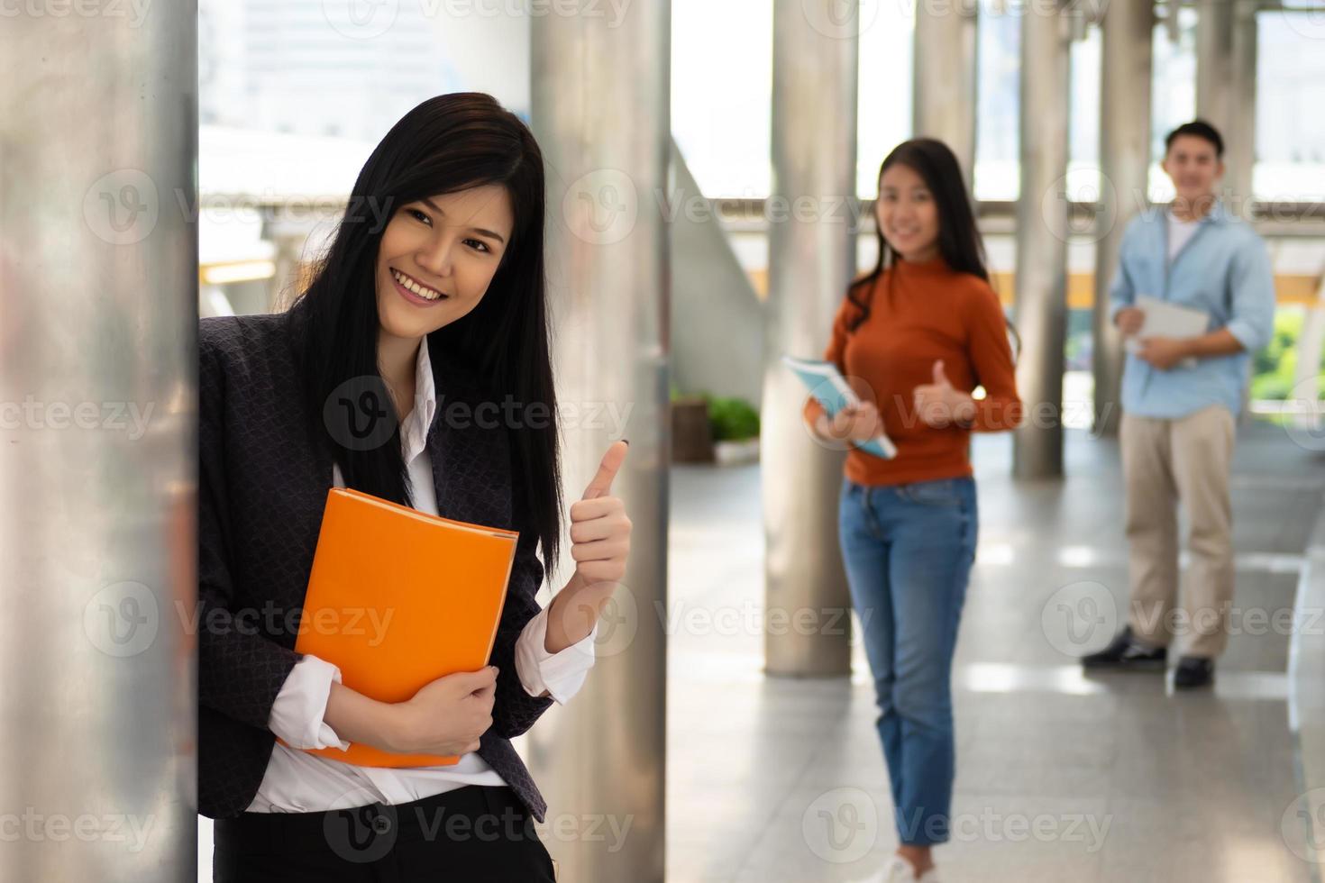 giovane asiatico donna Tenere libri e sorridente sollevato pollici su nel il Università, persone formazione scolastica concetto. foto