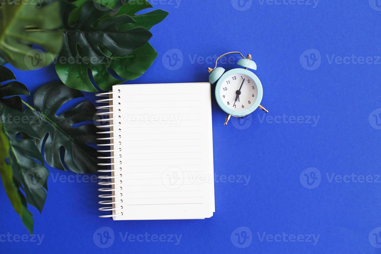 superiore Visualizza di vuoto Nota carta, allarme orologio e Monstera foglia su blu sfondo foto
