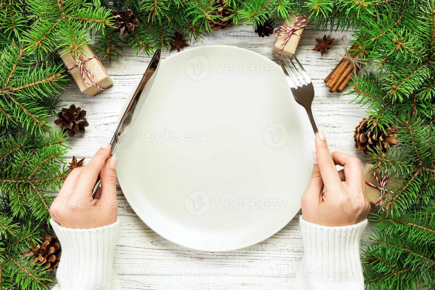 superiore Visualizza ragazza detiene forchetta e coltello nel mano e è pronto per mangiare. vuoto piatto il giro ceramica su di legno Natale sfondo. vacanza cena piatto concetto con nuovo anno arredamento foto