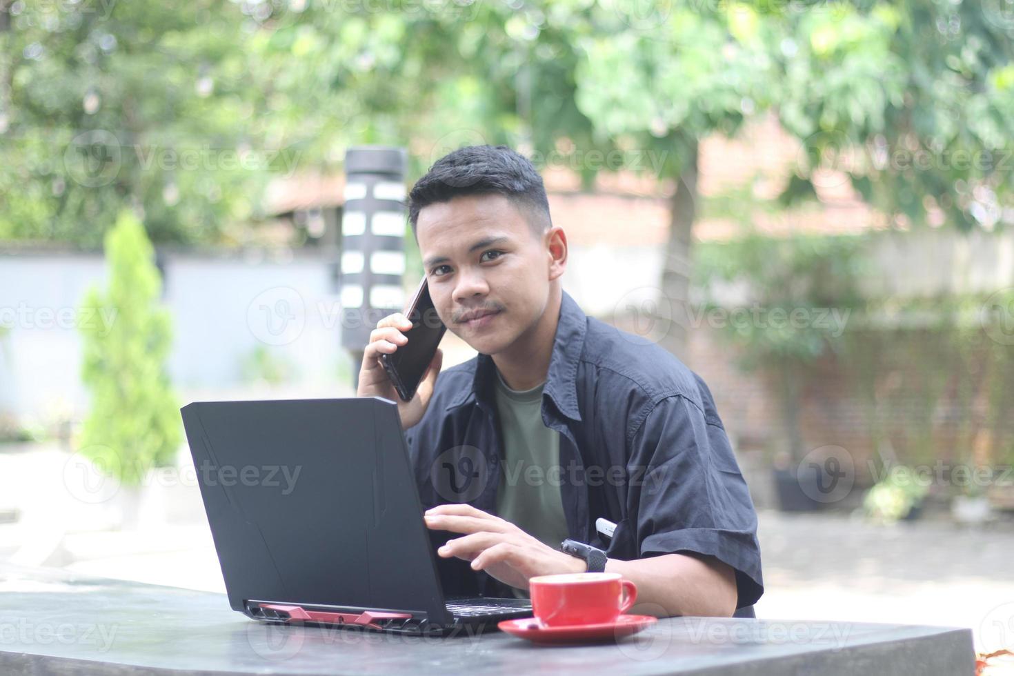 attraente giovane asiatico uomo utilizzando il computer portatile nel co-working spazio con contento viso e chiamata sua amico foto