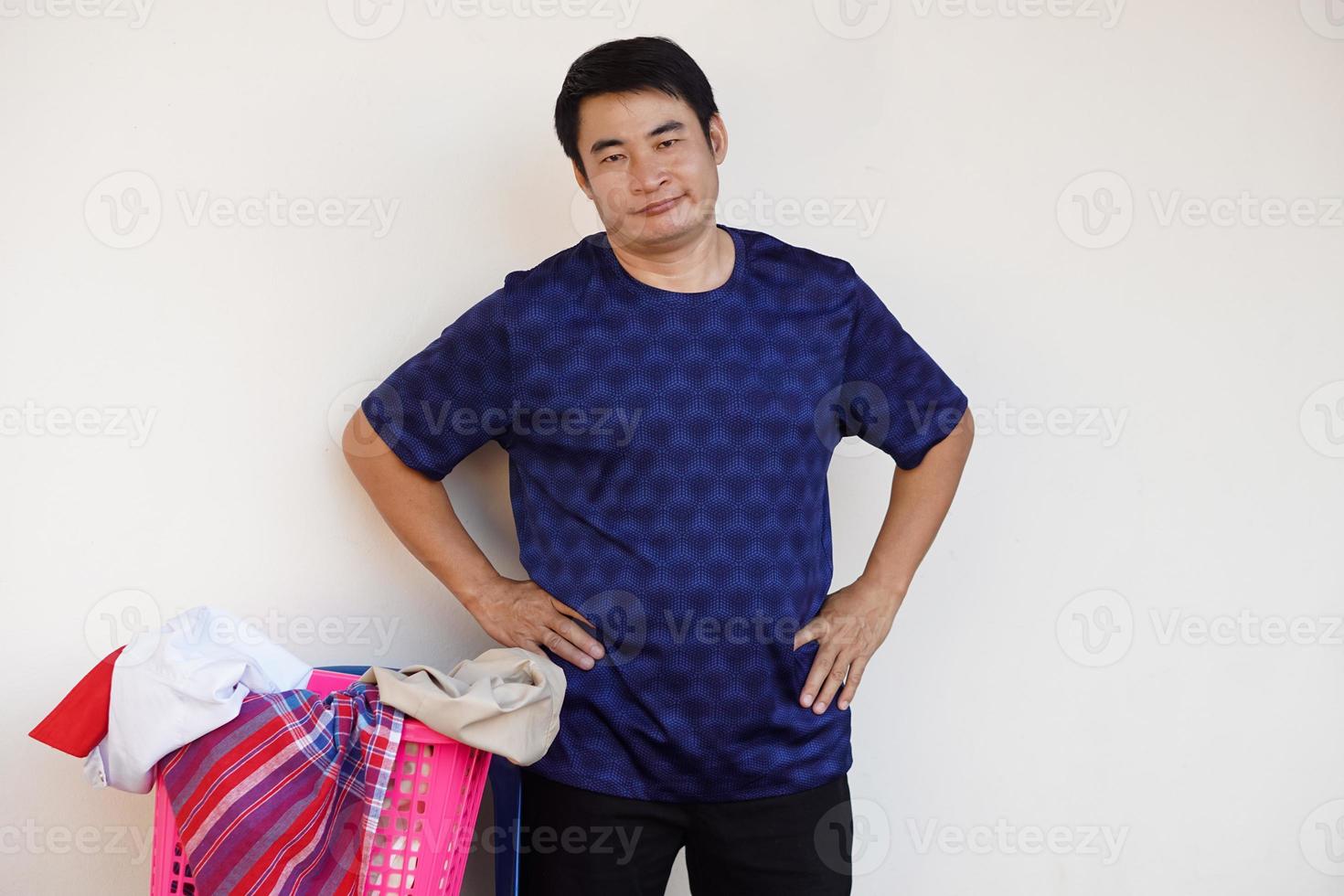asiatico uomo sta accanto cestino di Abiti, mettere mani su fianchi, sensazione quello lui non lo fa volere per fare lavanderia. concetto , noioso lavoretto ,lavori di casa. foto