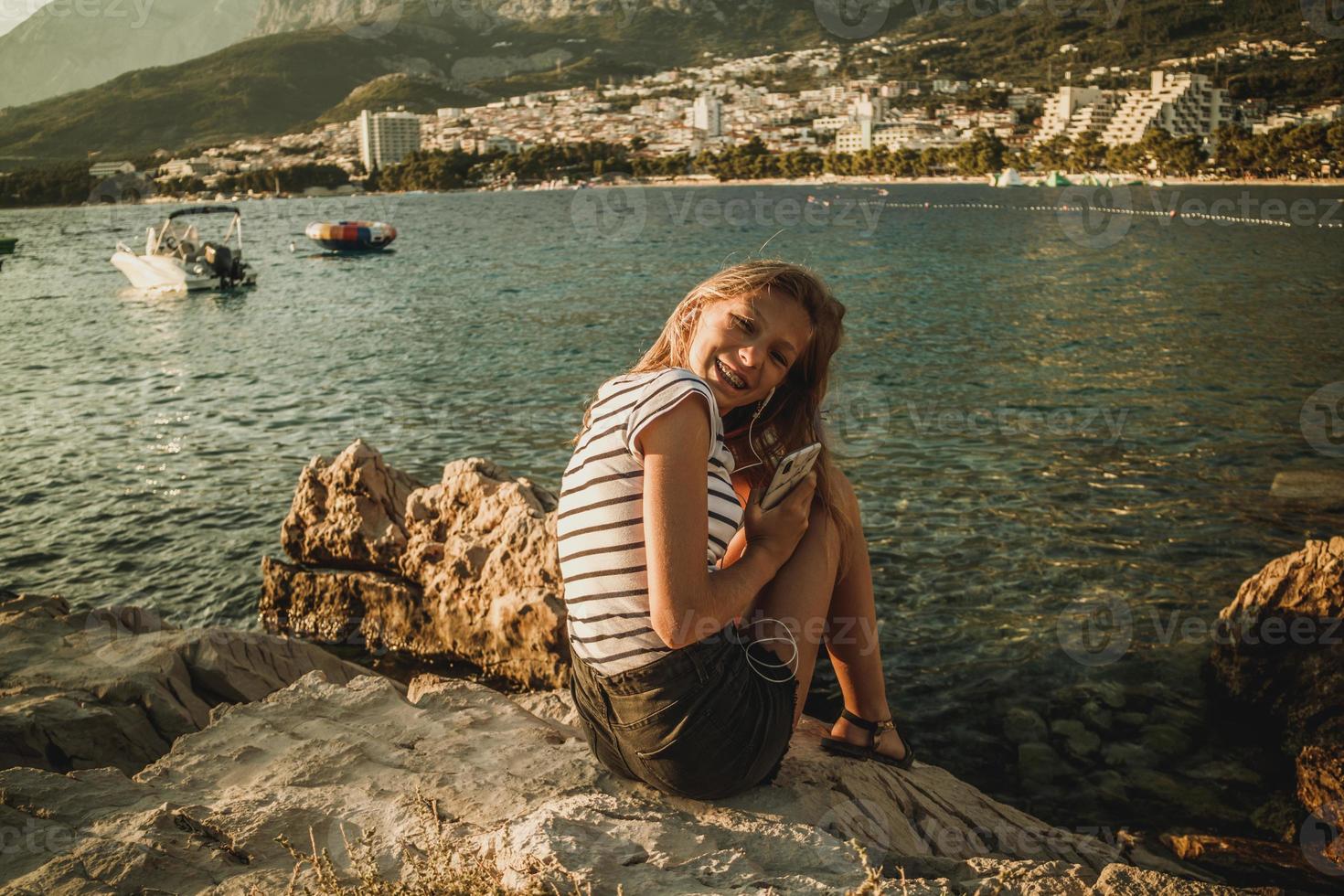adolescente ragazza seduta su un' roccia di il mare e godendo tramonto foto