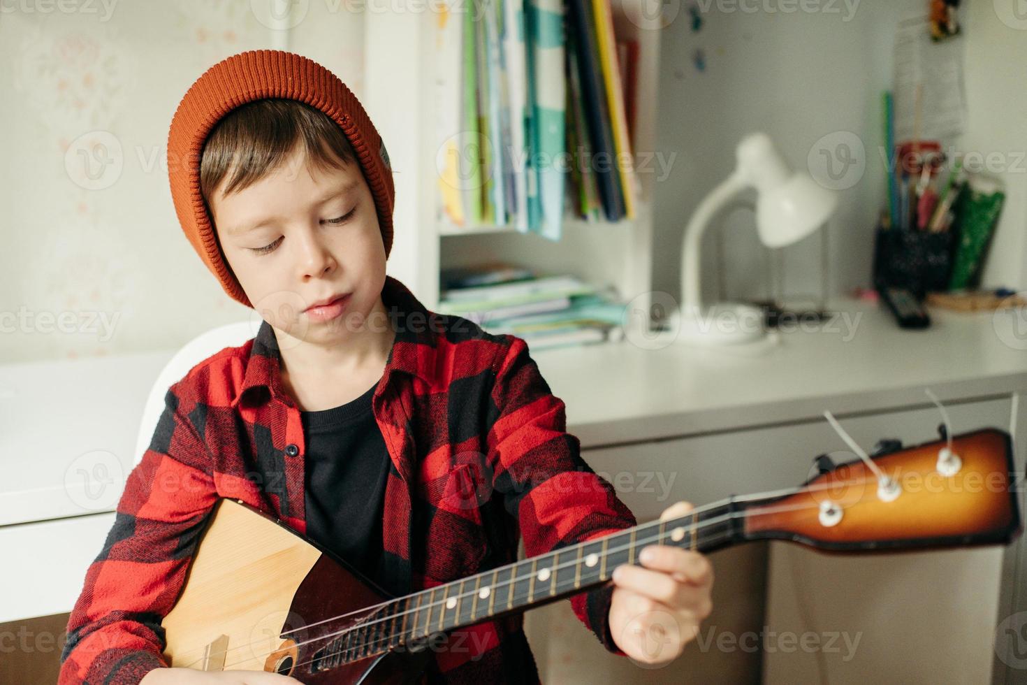 ragazzo nel un' rosso cappello e un' plaid camicia giochi il balalaica. bello ragazzo Tenere il suo chitarra. musica Lezioni a casa. passatempo per il anima. casa insegnamento musica foto