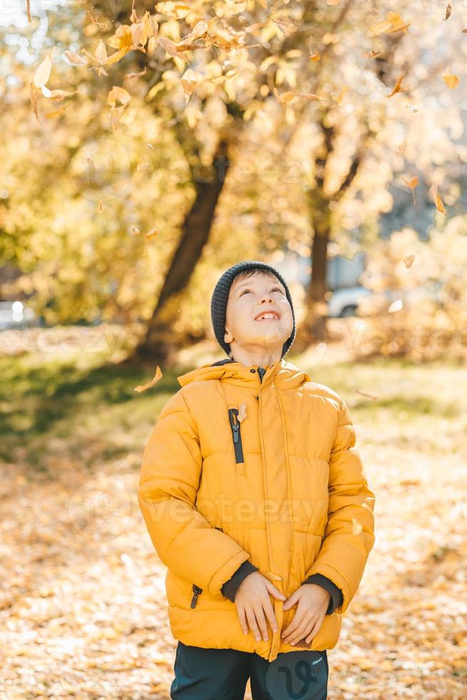 ragazzo nel un' giallo giacca, disperde le foglie nel un autunno parco. il bambino gioisce nel il autunno le foglie. contento infanzia. luminosa giallo giacca e le foglie foto