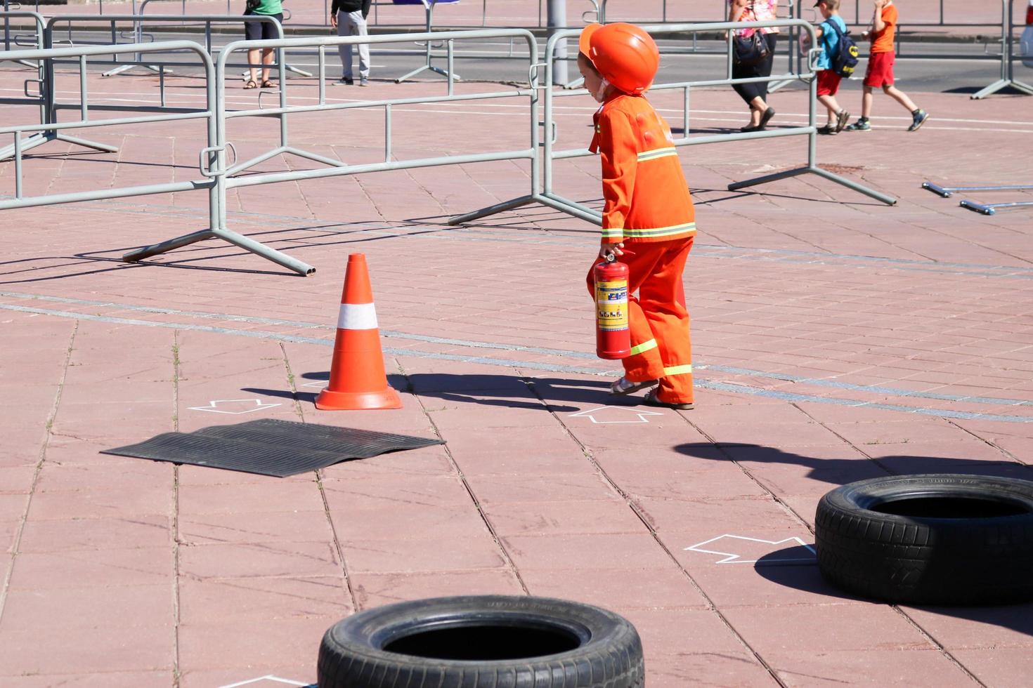 un' poco ragazza nel un scontroso resistente al fuoco completo da uomo corre con un' fuoco estintore per spegnere un' fuoco a un' concorrenza foto