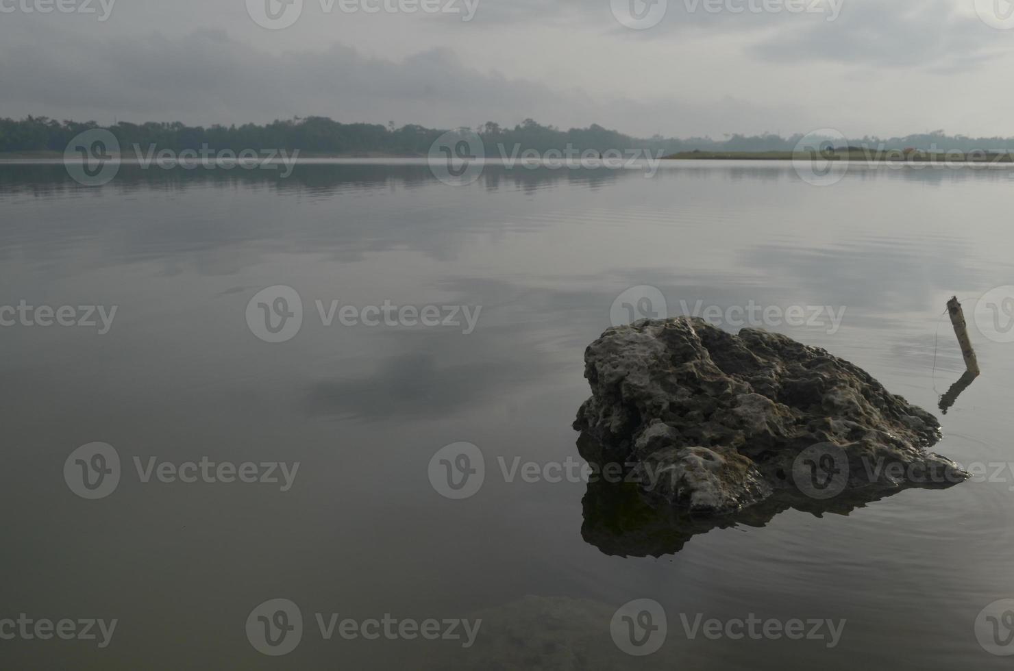 davanti Visualizza di un' grande roccia nel karangkate lago Indonesia con calma acqua condizioni a Alba con un' montagne sfondo e un' nebbioso cielo foto