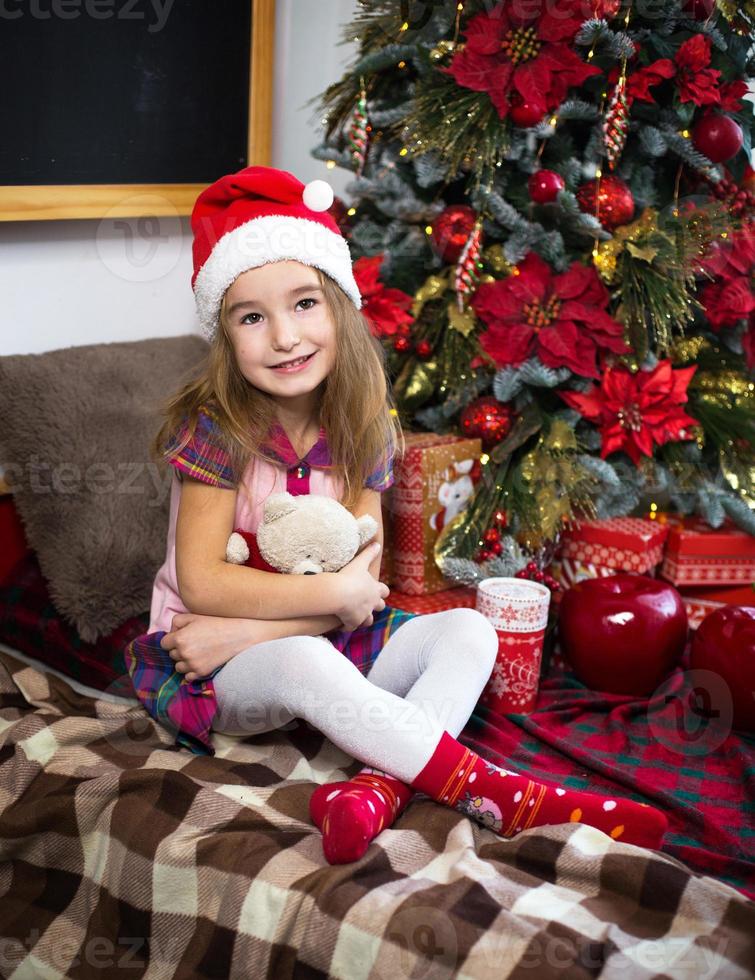 una bambina con in mano un orsacchiotto, seduta su una coperta a quadri nelle decorazioni natalizie vicino a un albero di natale con scatole di regali e un cappello da babbo natale. capodanno, gioco per bambini foto