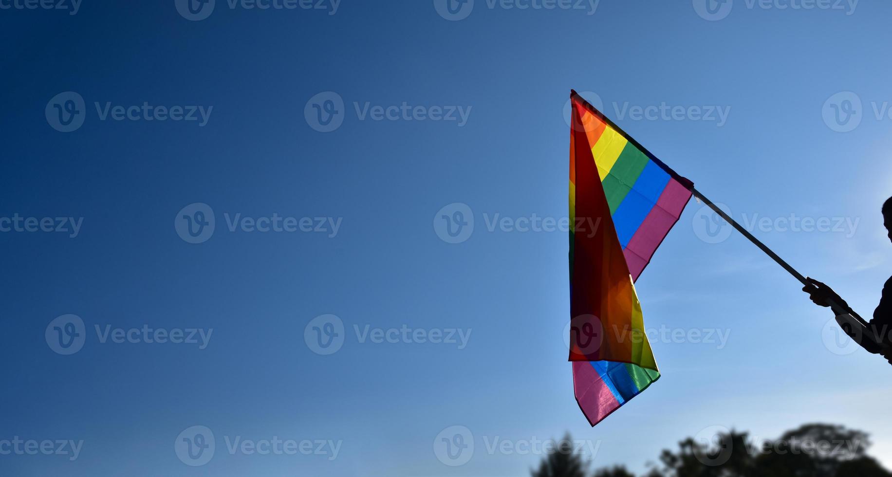 arcobaleno bandiera Tenere nel mano contro cielo blu sfondo, concetto per lgbt celebrazione nel orgoglio mese, giugno, in giro il mondo. foto