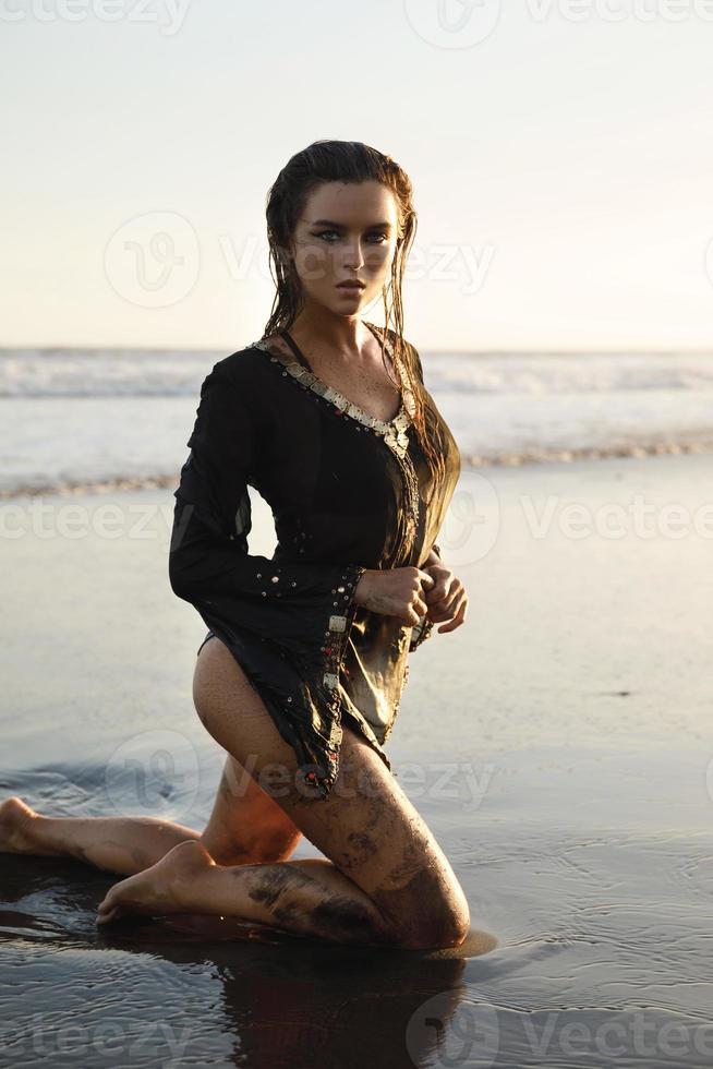 sexy donna indossare nero tunica è in posa su il spiaggia con nero sabbia foto