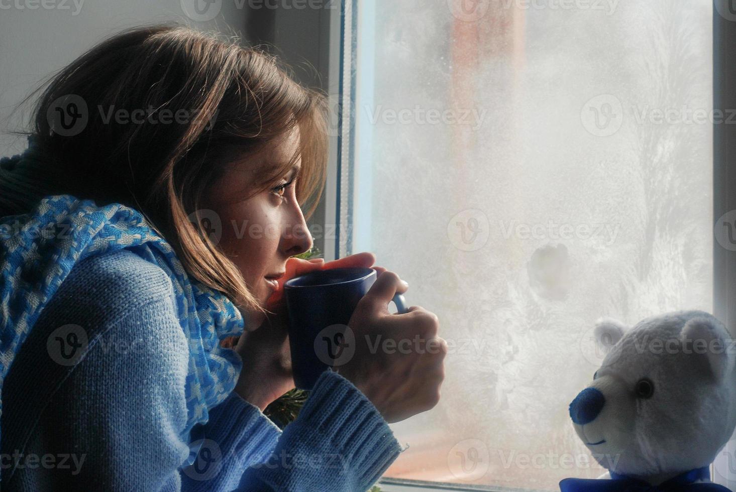 triste bella giovane donna soggiorno casa perché di il influenza e temperatura nel caldo Maglione e sciarpa con tazza di tè vicino il finestra foto