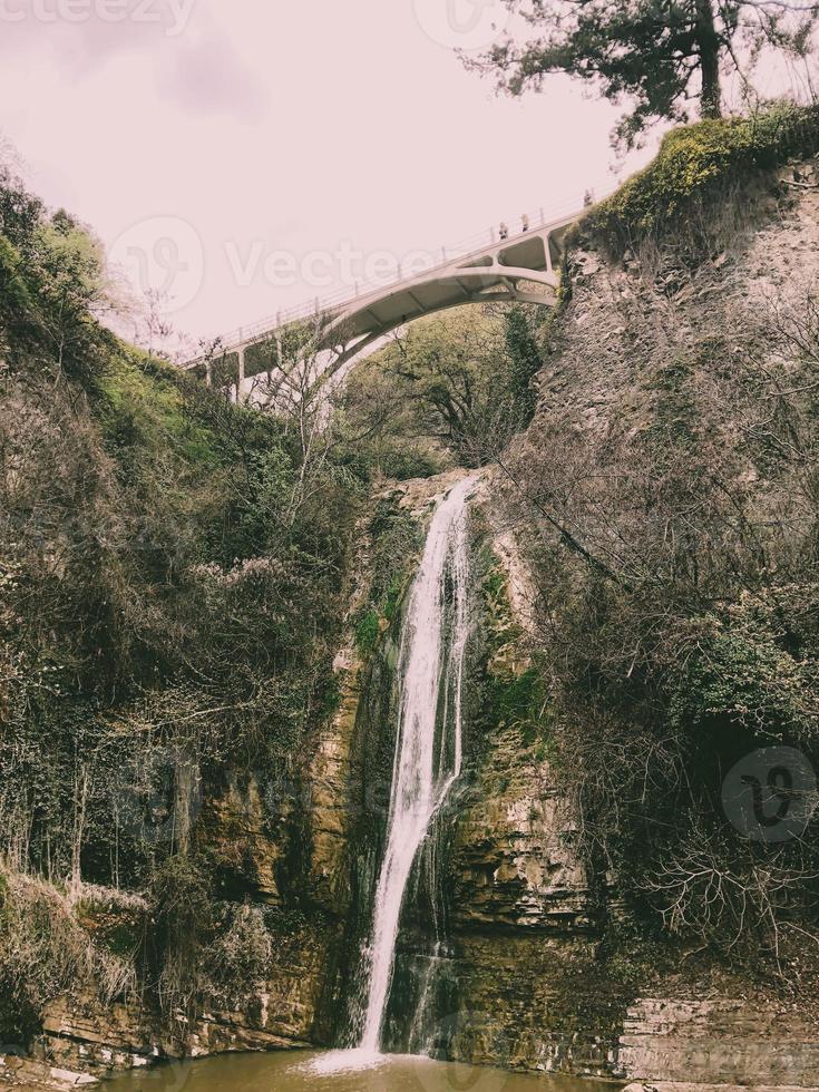 bellissimo montagna cascata con un' caduta ruscello di acqua fluente a partire dal sotto un' ponte su un' scogliera coperto di vegetazione con alberi e impianti foto